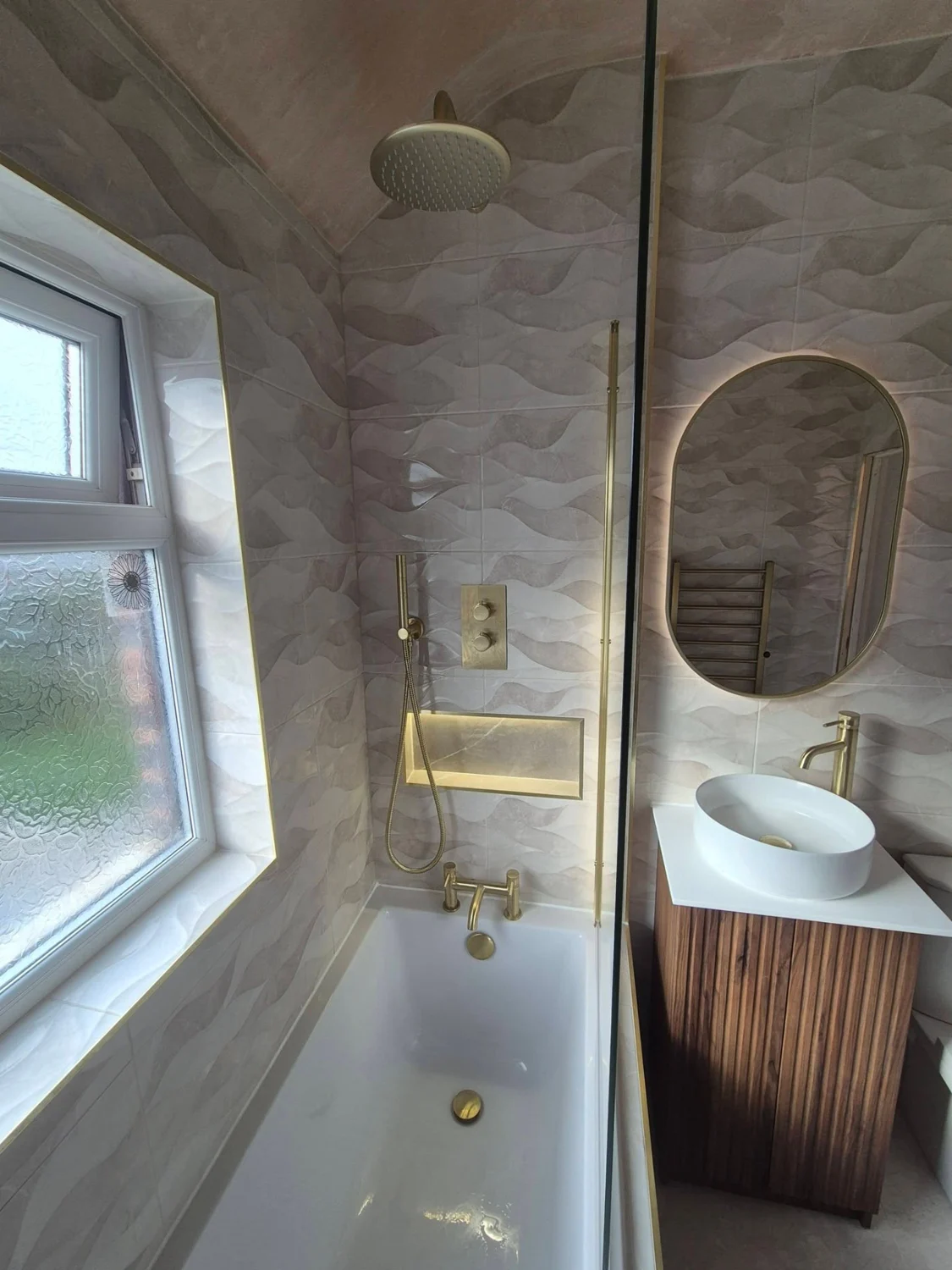 Completed bathroom with white bath, brass shower fittings, textured wall tiles and wood vanity